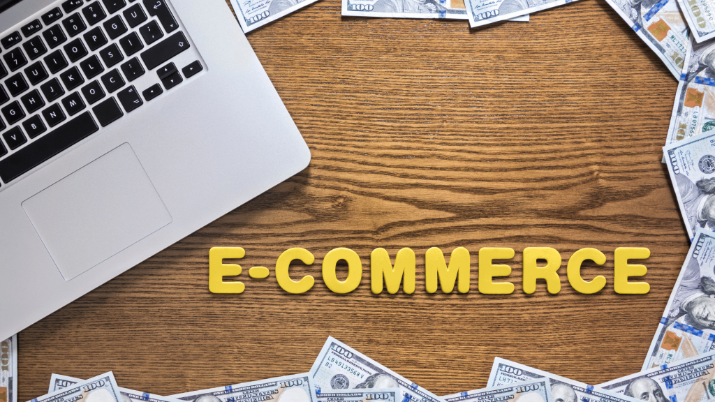 Flat lay of a laptop and scattered $100 bills on a wooden table with the text 'Why Pinterest Is a Hidden Gem for Ecommerce' over the word 'E-COMMERCE' spelled in yellow letters.